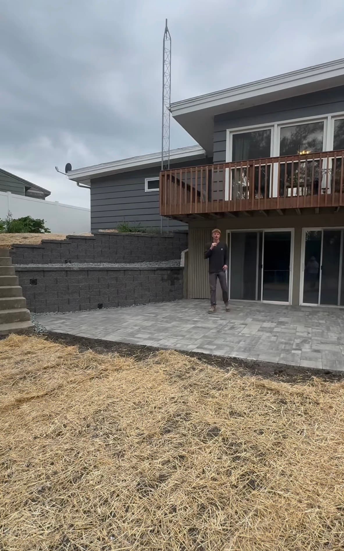 Man on a paved patio behind a house with a balcony and tiered retaining wall.