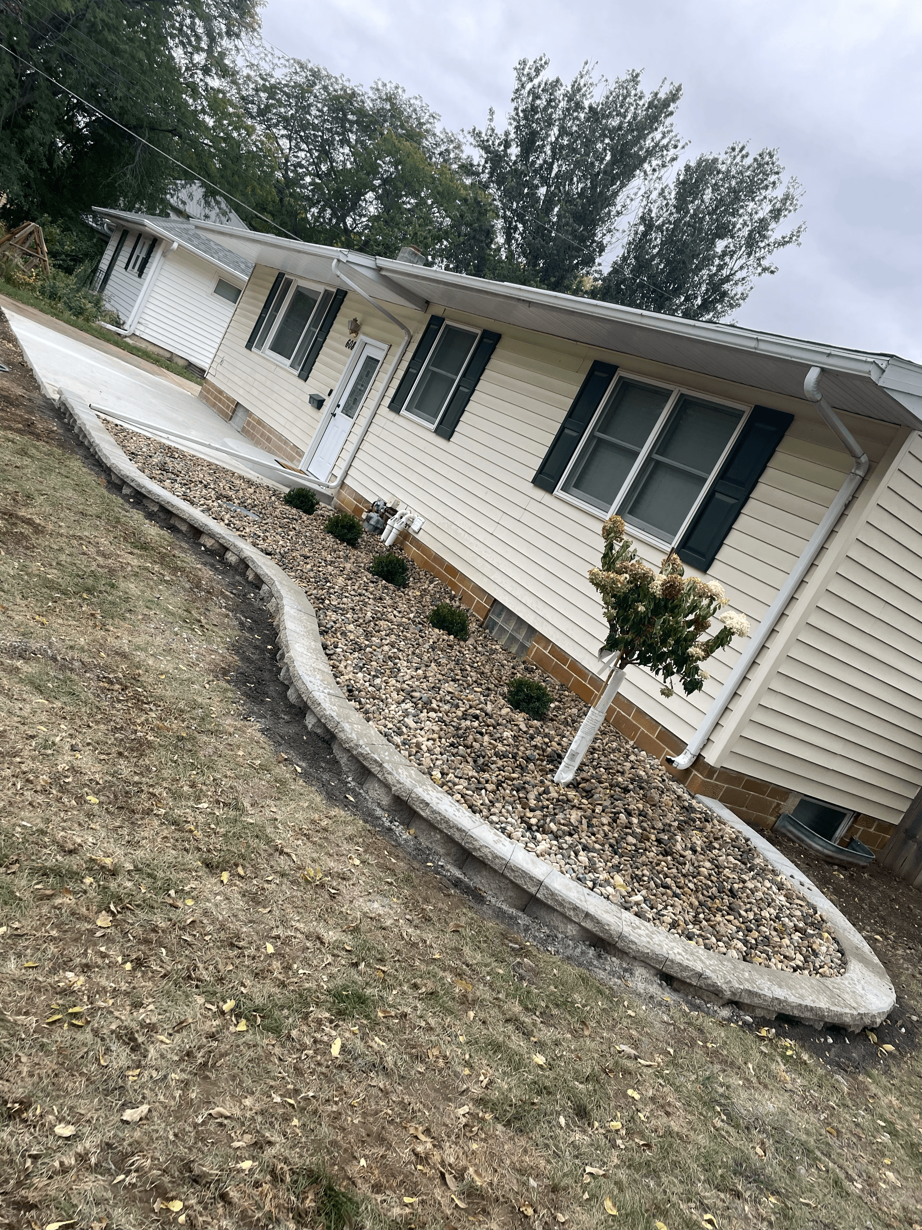 Cream house with dark shutters and a curved rock garden bed featuring a small tree.