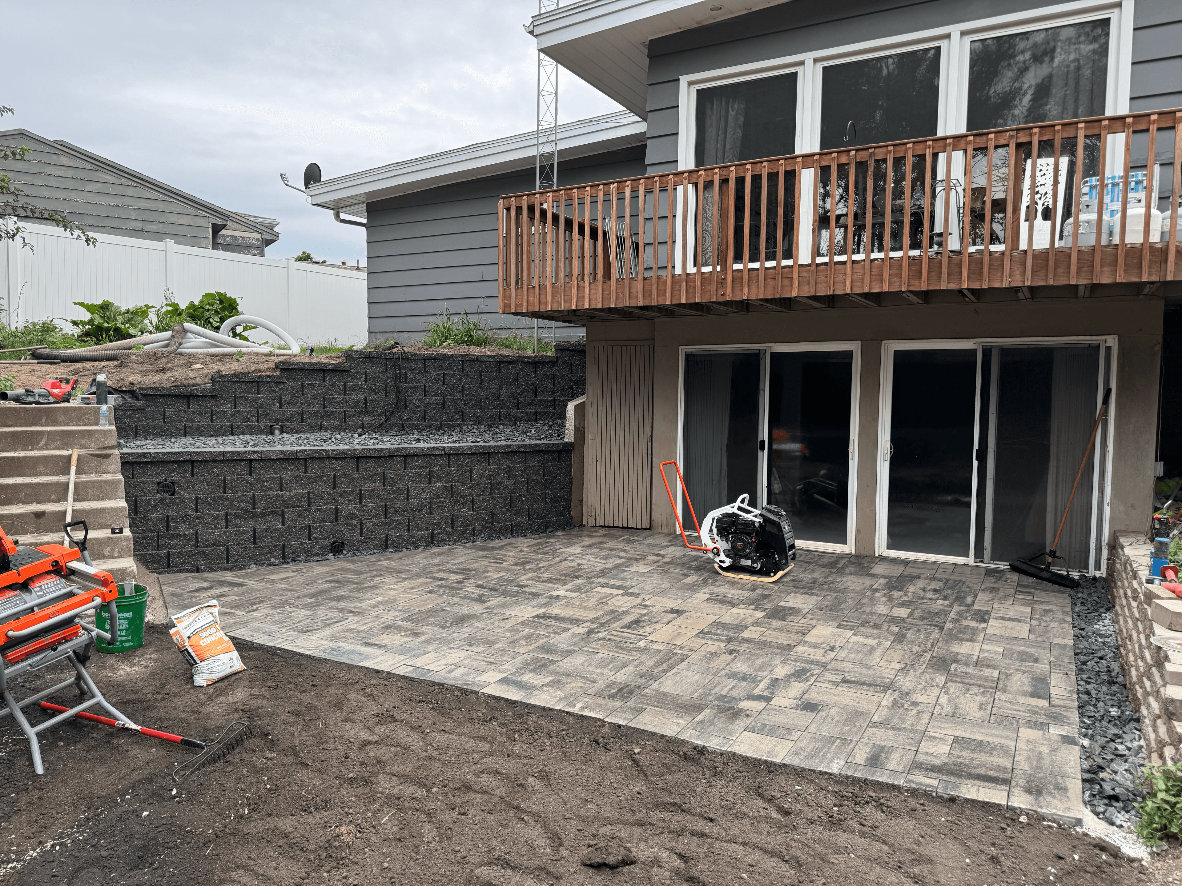 Newly laid grey stone paver patio with a plate compactor and tiered retaining wall.