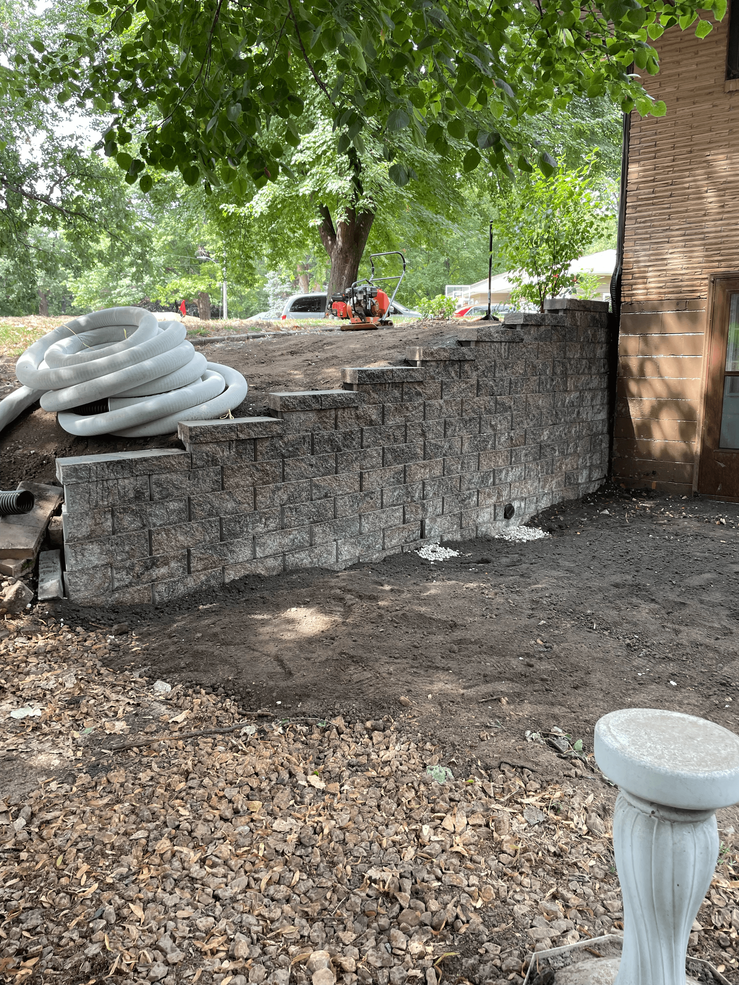 Tiered stone retaining wall under construction in a yard with coiled white drainage pipe.
