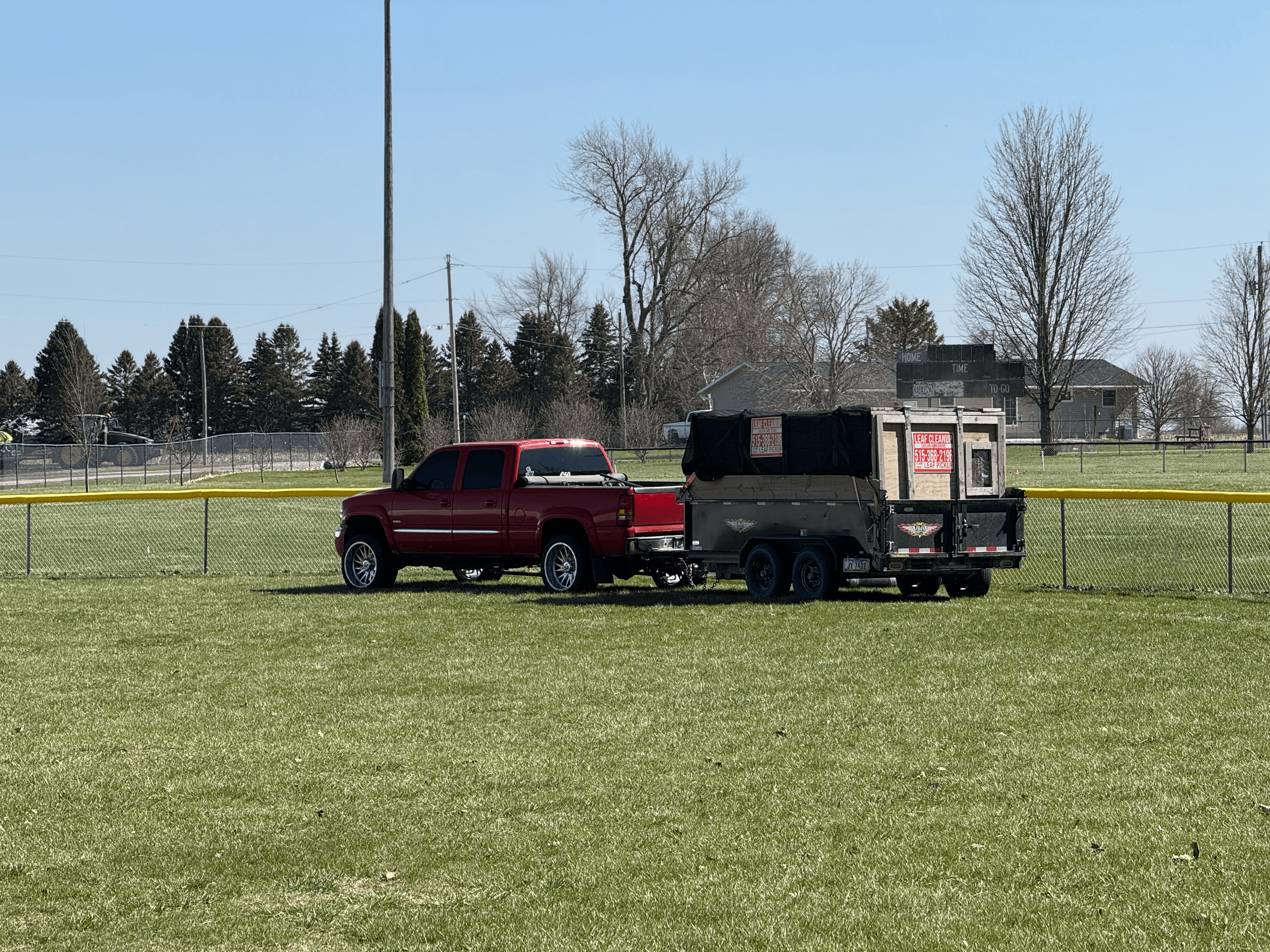 Red pickup truck towing a leaf cleanup trailer on a grassy field near a fence.